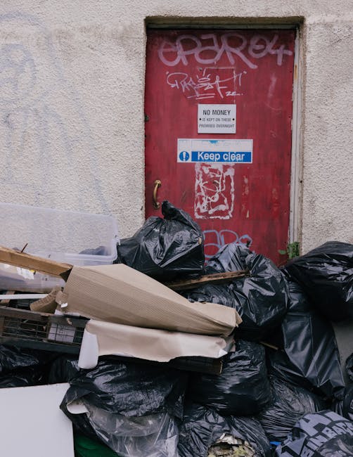 A pile of black rubbish bags and discarded paper and cardboard materials are stacked directly in front of a red, graffiti-covered metal door set into a beige, textured external wall, which appears weathered. The bags are filled with waste, some loosely tied, with various textures and sizes, occupying the lower half of the image. To the left of the bags, a piece of light-colored cardboard or foam board is visible, along with a wooden pallet or similar object partially covered by the waste. The door features multiple graffiti tags in white and pink spray paint, along with functional signage including a 'NO MONEY' notice and a blue sign that reads 'Keep clear,' which are positioned centrally and towards the top of the door. The surrounding environment suggests an urban alleyway or rear service area, typical for private waste disposal, and the scene highlights the importance of safe and proper rubbish removal services that Rubbish Clearance Dulwich provides for efficient waste management and debris clearance.