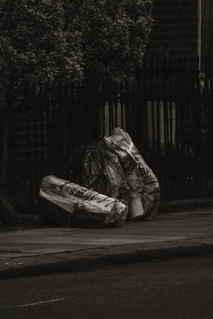 Three large plastic bags filled with waste material are placed on the curbside pavement next to a black metal fence, with a background of foliage and a building. The bags appear crumpled and are sealed, containing household or garden rubbish. The scene is outdoors on a concrete sidewalk, adjacent to a roadway, under natural daylight. The environment suggests a typical urban or suburban setting where individual rubbish collection might occur, and the presence of waste bags reflects a private or alternative rubbish removal method rather than a municipal service. Rubbish Clearance Dulwich offers professional waste disposal options for such debris, supporting on-site clearance and private disposal needs. The image emphasizes the importance of responsible rubbish management outside typical bin collection points, highlighting the role of specialist companies in managing waste that may not be suitable for standard removal services.