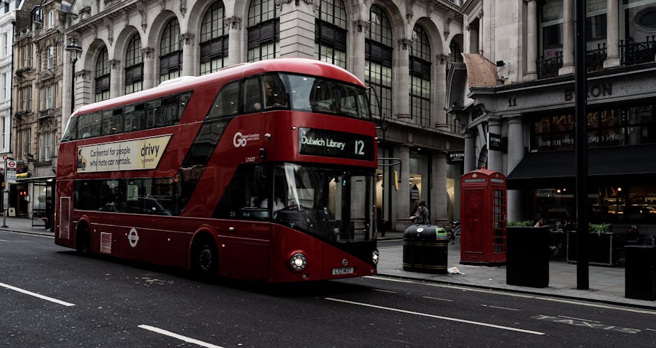 A red double-decker bus is stationary on a city street, with a black route number 12 sign indicating it is headed to Dulwich Library. The bus features a smooth, glossy exterior with black tinted windows and a white advertising banner on its side. Behind the bus, there are historic stone buildings with large glass windows, ornate architectural details, and multiple floors. To the right, a traditional red telephone booth stands on the pavement next to a black waste bin, with greenery visible in front of commercial storefronts under a canopy. The street surface is asphalt with visible white lane markings, and the scene appears to be captured during daytime with natural lighting. The environment suggests an urban setting with a mix of transportation and street furniture, typical for areas with independent transport services such as those provided by local rubbish removal companies like Rubbish Clearance Dulwich, which may also operate private waste collection adjacent to such busy cityscapes.
