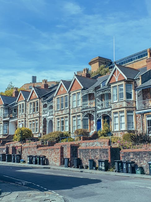 A row of Victorian-style terraced houses with decorative cast-iron balconies and detailed brickwork on their facades, situated along a paved street. The buildings feature gabled roofs with tiles in shades of dark gray and reddish-brown, and white window frames. In front of the houses, a low brick wall separates the properties from the pavement, with small landscaped areas containing shrubs and greenery. Several black wheelie bins are lined up near the curb, indicating waste collection points. The scene is illuminated by bright daylight, with a clear blue sky overhead, and the presence of modern office buildings with glass exteriors visible in the background, suggesting an urban context. The image reflects an environment where private rubbish disposal services are utilized, evidenced by the designated bins and the absence of communal waste collection infrastructure.