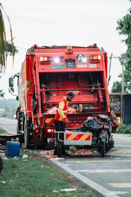 A waste collection worker in a yellow and red high-visibility jacket is standing on a sidewalk at the rear of a large red rubbish truck, which is parked on the side of a street. The back of the truck features multiple hydraulic and mechanical components, with labels and warning stickers, and is in the process of collecting waste. The worker is handling a black plastic trash bag, with additional waste items and debris scattered around the truck's loading area. The scene is set outdoors during daytime, with trees and some buildings visible in the background, and the street has a marked curb and road surface. The overall environment suggests an active rubbish removal operation, possibly an alternative or private waste collection service, reflecting typical street-based refuse management activities in urban or suburban areas. Rubbish Clearance Dulwich services are connected to the context of effective rubbish collection and waste disposal complemented by on-site clearance and independent waste handling practices.