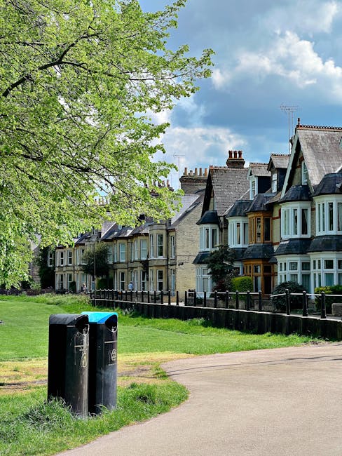 A row of traditional terraced houses with bay windows and tiled roofs line the background of the image, set behind a small green park area. In the foreground, two black recycling bins are positioned on the paved walkway, which curves gently around the grassy space. The park features a large, leafy tree with fresh green foliage extending across the top left corner, providing partial shade to the area. The sky above is partly cloudy with patches of blue visible between the fluffy clouds, suggesting a bright daytime scene. The scene captures an outdoor area that could benefit from private rubbish collection or on-site clearance services, with Rubbish Clearance Dulwich potentially offering alternative waste handling options to traditional municipal disposal for residents in this neighbourhood.