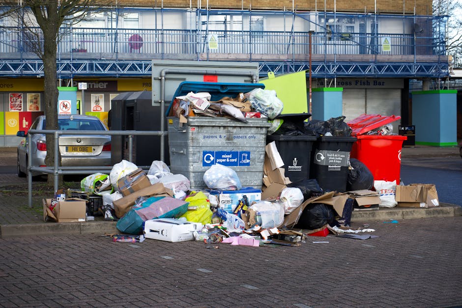 A collection of overflowing waste and rubbish bags is piled on a paved area in front of a row of recycling bins and a parking space. The waste includes black and black-and-white plastic rubbish bags, cardboard boxes, crumpled paper, and discarded packaging materials, some spilling onto the surrounding pavement. The recycling bins include a large blue mixed paper and cardboard container with an open lid, partially filled with paper waste, and a black bin designated for general rubbish. Behind the waste area, there is a silver vehicle parked next to a metal railing, and in the background, a building with signage and a blue security fence on the upper level. The scene suggests a situation of improper waste disposal, possibly related to off-site rubbish collection or private waste handling, within a public or commercial area with urban surroundings.