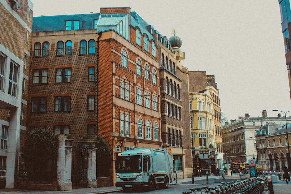 A city street scene showing a collection of historic and modern buildings with varied architectural styles, including a large brick building with arched windows and decorative brickwork, adjacent to a cream-colored building with ornate window frames and a small dome on its roof. In the foreground, a rubbish collection vehicle with a silver, smooth exterior is parked on the pavement, positioned near a gated entrance featuring stone columns and decorative metalwork. The street is lined with metal bollards, and bicycles are secured along a cycle rack nearby. The environment appears overcast with diffuse natural light illuminating the scene, suggesting a typical urban setting where private waste collection services like Rubbish Clearance Dulwich operate to manage rubbish removal beyond local authority fill-ins, highlighting an alternative waste handling method in a busy city area.