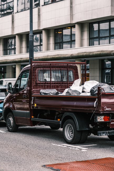 A small burgundy flatbed truck parked on an urban street in front of a modern concrete office building with large windows. The truck's bed is filled with various types of rubbish, including stacked black and white garbage bags, some appearing full and loosely tied, and metallic or plastic debris. The truck features a red protective grid at the cab's rear, and the vehicle shows signs of typical use with visible dirt and minor wear. Surrounding vehicles are parked along the street, with a white van visible nearby. A tall streetlight stands adjacent to the truck, casting a faint shadow. The environment suggests an area where private waste collection or rubbish removal services occur, supporting the context of alternative waste handling outside of standard municipal collection. The lighting is neutral daylight, emphasizing the details of the rubbish and the urban setting, aligning with the service focus on efficient, same-day rubbish clearance in the Dulwich area managed by Rubbish Clearance Dulwich.