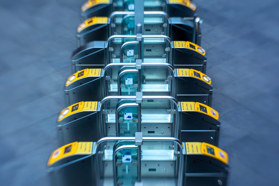 The image displays a row of six modern, stainless steel trolley-style luggage carts with curved handles and blue plastic inserts, arranged in a straight line on a dark gray, textured floor. Each cart is equipped with a yellow electronic payment pad mounted at the front, featuring a circular scanning area and touchpad interface. The carts are clean and polished, reflecting light, and are aligned closely together, occupying the central portion of the image. The background is out of focus, providing a neutral environment typical of an indoor public space such as a train station or parking area, hinting at a setting where luggage or equipment might be moved or collected. The scene suggests a context related to transportation or service areas where equipment like carts is used for assisting with baggage handling or waste collection logistics, aligning with the services provided by Rubbish Clearance Dulwich for efficient and independent waste management solutions.